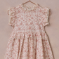 Noralee's Baby Girls' and Girls' Alice Dress features a pink floral pattern, ruffle sleeves, and an embroidered lace neckline, showcased on a copper hanger against a textured pink background.