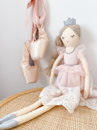 The Mon Ami Princess Olivia Doll, dressed in a pink lace dress and crown, sits on a wicker surface next to a pair of pink ballet shoes hanging on the wall, making her a delightful keepsake gift for any ballet lover.