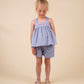 A young child stands facing forward, wearing the Early Sunday Girls’ Roman Shorts paired with a light blue sleeveless top adorned with pink hearts and light-colored sandals, set against a beige background.