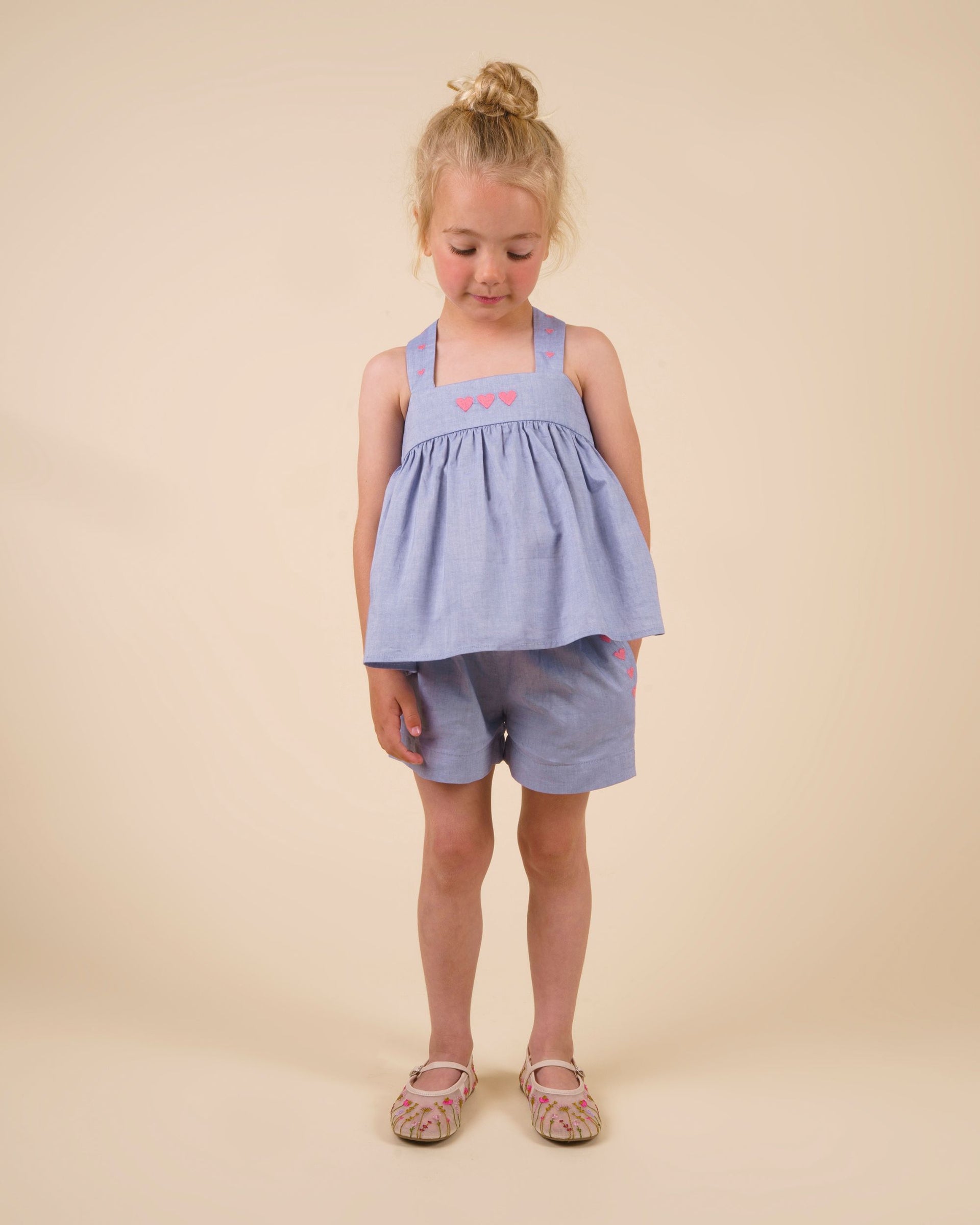 A young child stands facing forward, wearing the Early Sunday Girls’ Roman Shorts paired with a light blue sleeveless top adorned with pink hearts and light-colored sandals, set against a beige background.