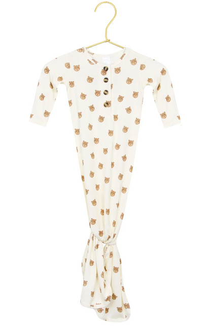 The Lou Lou & Company Baby Teddy Knotted Gown, featuring a cream color, long sleeves, wooden buttons, and an adorable bear face print, hangs on a gold hanger. This tie-bottom gown is designed with a knotted bottom for easy diaper changes and includes a convenient button-down front.