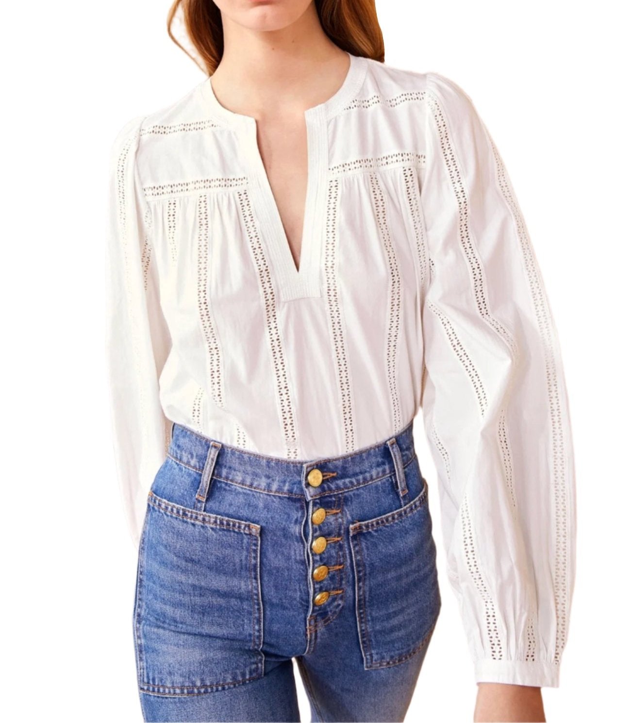 A person dressed in the Ulla Johnson Rowan Blouse featuring geometric eyelets and high-waisted blue jeans adorned with gold buttons is standing in front of a textured wall.