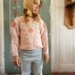 A young child stands indoors against a painted door and white wooden wall, wearing the Louise Misha Girls’ Acacia Sweater—a pink knit piece featuring handmade embroidery—paired with a hat decorated with playful pom-poms and light blue pants.