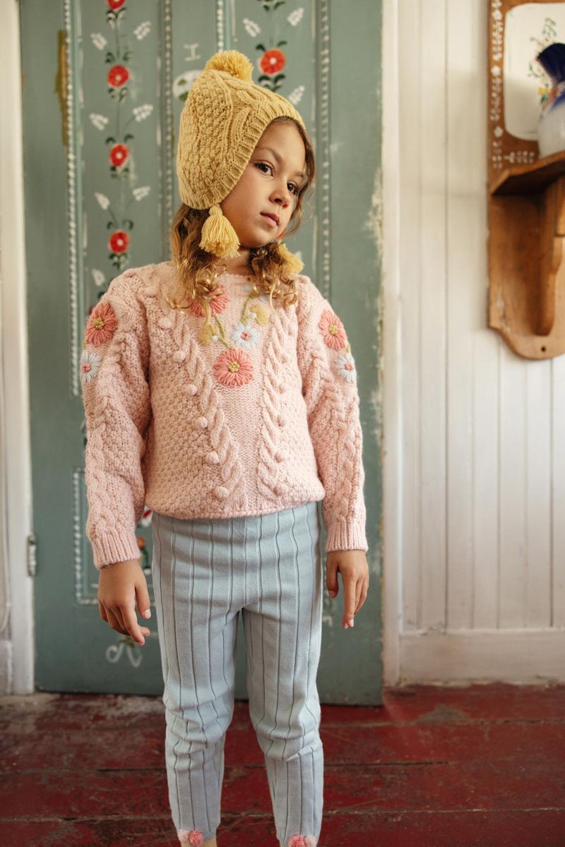 A young child stands indoors against a painted door and white wooden wall, wearing the Louise Misha Girls’ Acacia Sweater—a pink knit piece featuring handmade embroidery—paired with a hat decorated with playful pom-poms and light blue pants.
