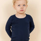A young boy with curly blonde hair and blue eyes wears the Copenhagen Colors Baby Boys’ and Boys’ Merino Long Sleeve Knitted T-Shirt, standing against a beige background.