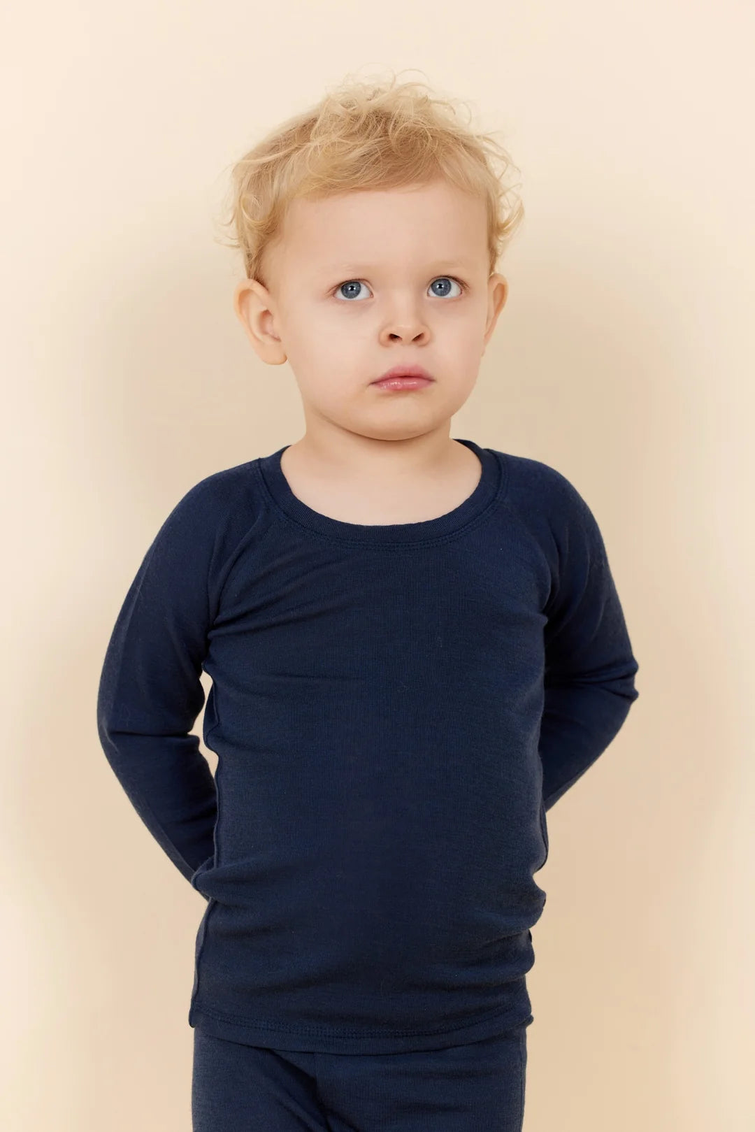 A young boy with curly blonde hair and blue eyes wears the Copenhagen Colors Baby Boys’ and Boys’ Merino Long Sleeve Knitted T-Shirt, standing against a beige background.