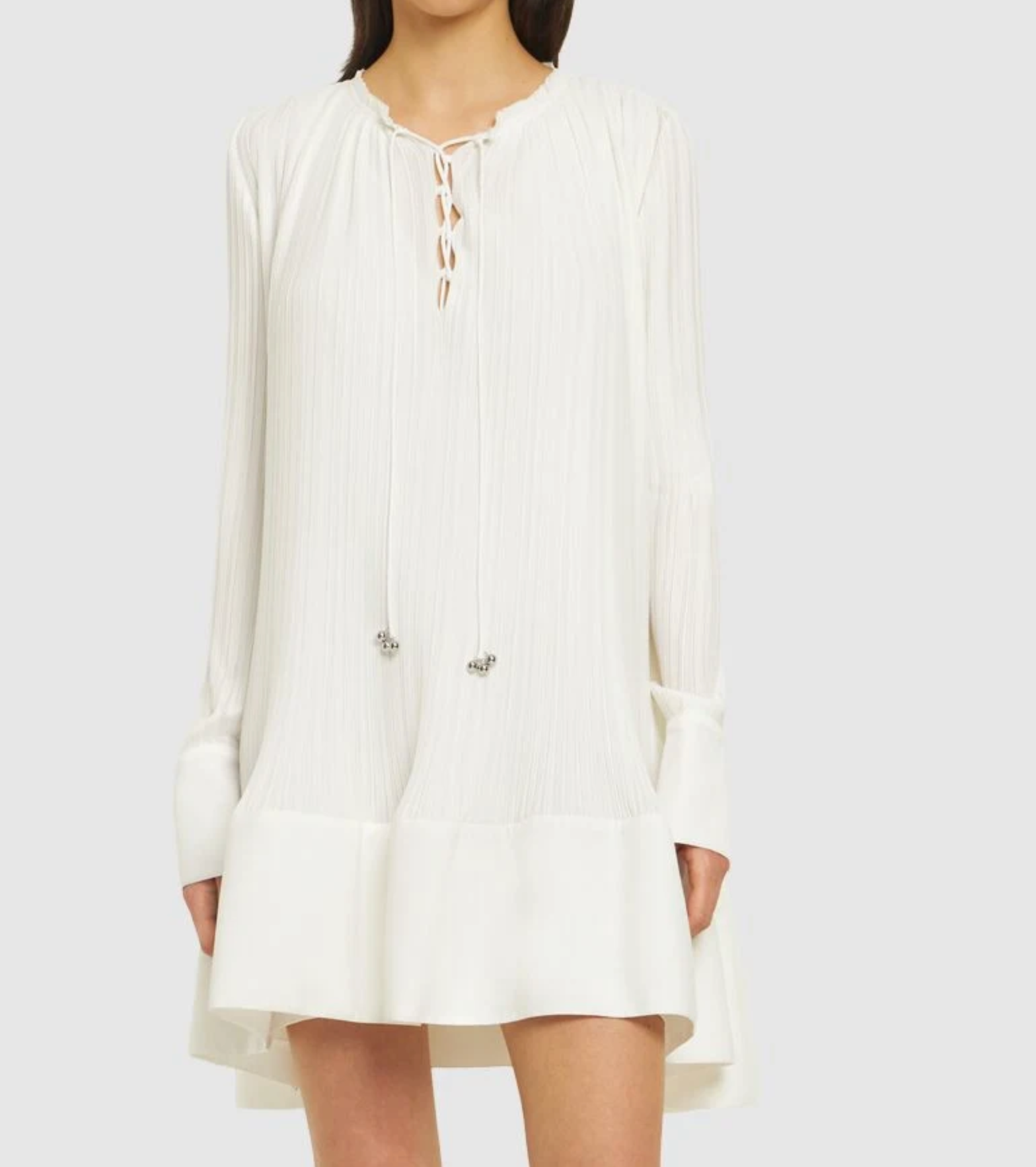 A woman models the Lanvin Flared Pleated Dress with Long Sleeves, a loose-fitting white mini dress featuring a lace-up neckline, ruffled hem, and delicate pleats, set against a plain light background.