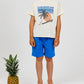 A barefoot child stands next to a pineapple, wearing a cream "Caribbean Tranquilo" T-shirt with a sunset print and bright blue Boys' Cotton Rustic Bermuda Shorts by Arsene & Les Pipelettes, featuring an elasticated waist and crafted from textured cotton, set against a plain light background.