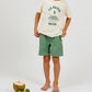 A child stands barefoot wearing a "La Pasta E Basta" T-shirt and Arsene & Les Pipelettes Boys' Cotton Rustic Bermuda Short, featuring an elasticated waist in green, next to a coconut with a straw and three limes on a white background.