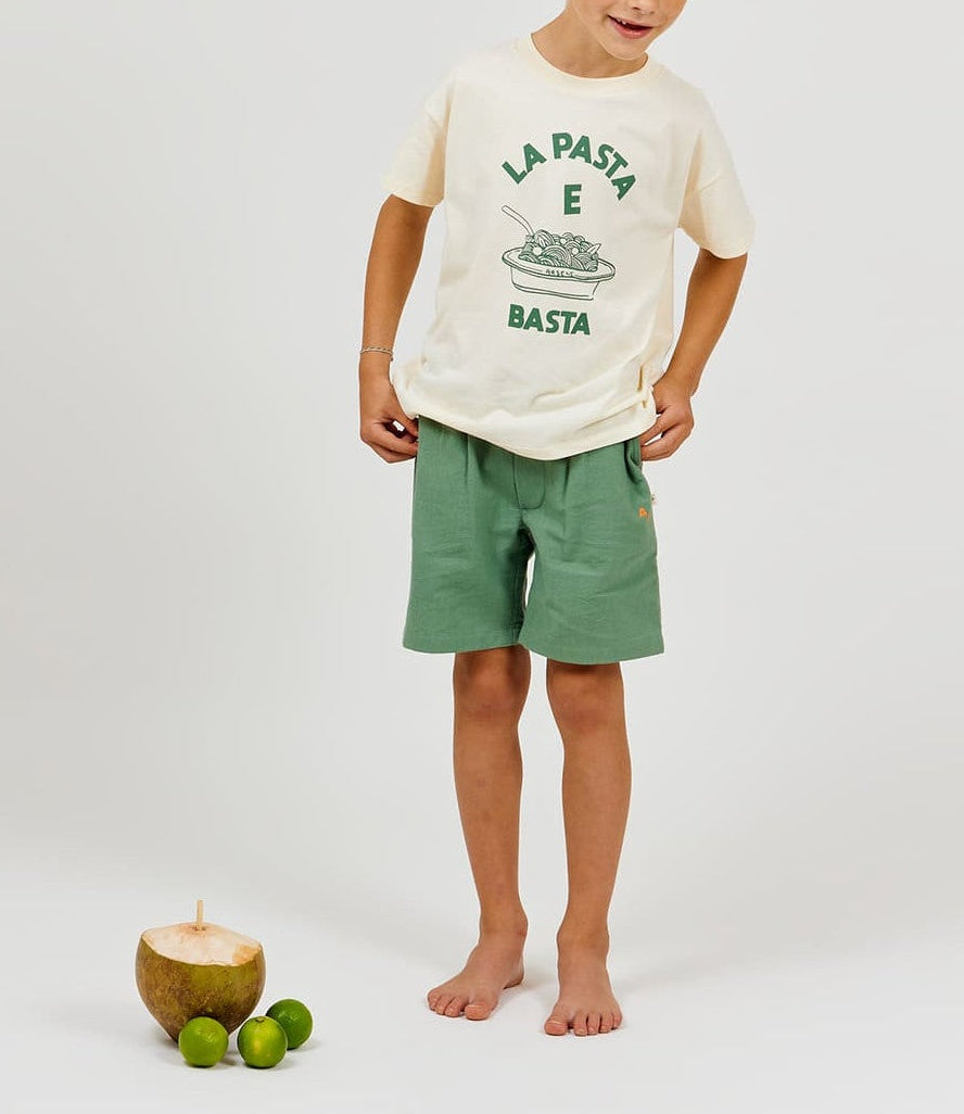 A child stands barefoot wearing a "La Pasta E Basta" T-shirt and Arsene & Les Pipelettes Boys' Cotton Rustic Bermuda Short, featuring an elasticated waist in green, next to a coconut with a straw and three limes on a white background.