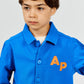A young boy wears the Arsene & Les Pipelettes Boys' and Girls' Cotton Rustic Jacket, featuring vintage-inspired blue fabric with bold orange "AP" letters on the chest, standing against a plain background.