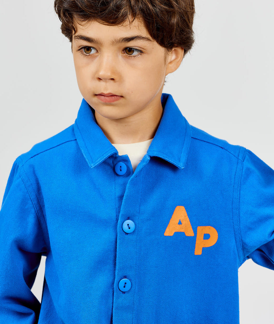 A young boy wears the Arsene & Les Pipelettes Boys' and Girls' Cotton Rustic Jacket, featuring vintage-inspired blue fabric with bold orange "AP" letters on the chest, standing against a plain background.