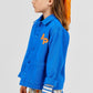 A young child wearing the Arsene & Les Pipelettes Boys' and Girls' Cotton Rustic Jacket in vintage-effect blue, styled with an orange scarf in their hair and a white skirt adorned with blue and orange patterns, stands in profile against a plain background.