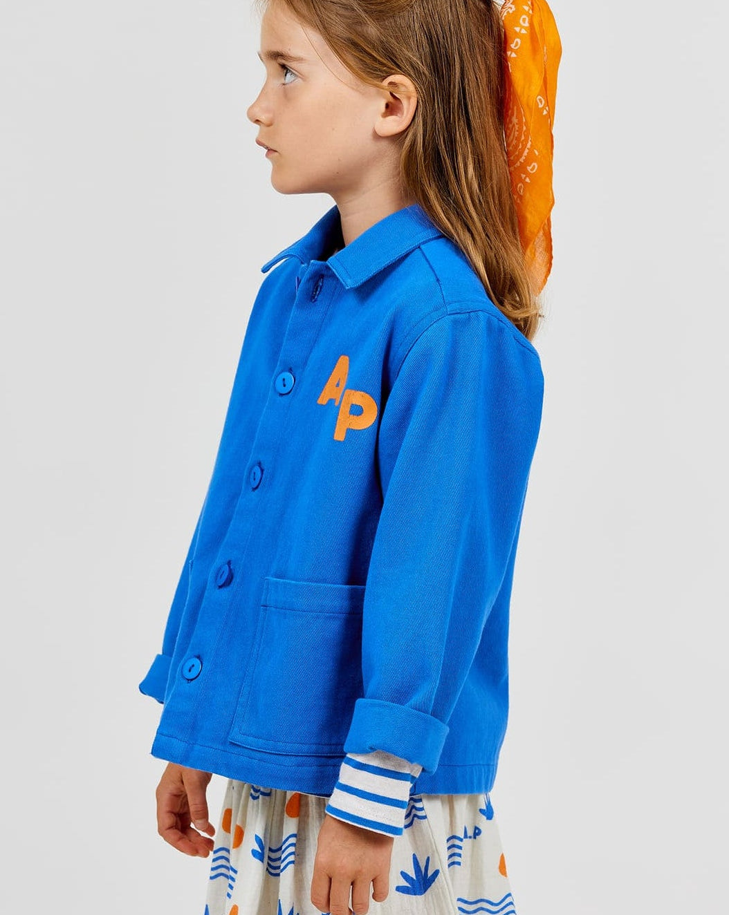 A young child wearing the Arsene & Les Pipelettes Boys' and Girls' Cotton Rustic Jacket in vintage-effect blue, styled with an orange scarf in their hair and a white skirt adorned with blue and orange patterns, stands in profile against a plain background.