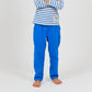A child stands barefoot with arms crossed, dressed in Arsene & Les Pipelettes' Boys' Cotton Rustic Pant in bright blue, featuring an elastic waist, paired with a long-sleeve white shirt adorned with blue stripes and small colorful chest designs, set against a plain background.