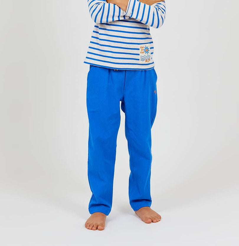 A child stands barefoot with arms crossed, dressed in Arsene & Les Pipelettes' Boys' Cotton Rustic Pant in bright blue, featuring an elastic waist, paired with a long-sleeve white shirt adorned with blue stripes and small colorful chest designs, set against a plain background.