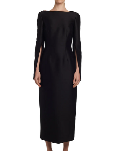 A person wears the Adam Lippes Talitha Silk Wool Twill Dress, a long black Japanese silk wool design with a high boat-neck and split sleeves, standing against a white background.