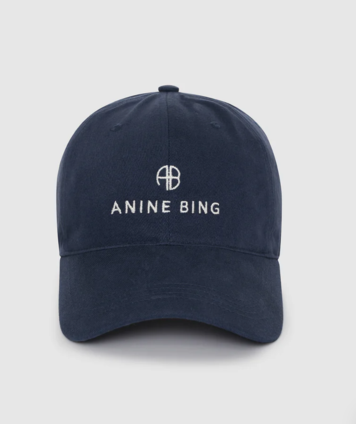 The Jeremy Baseball Cap by Anine Bing is a navy blue cotton-twill hat with "ANINE BING" and a circular AB logo embroidered in white on the front, plus an adjustable back strap for a custom fit.