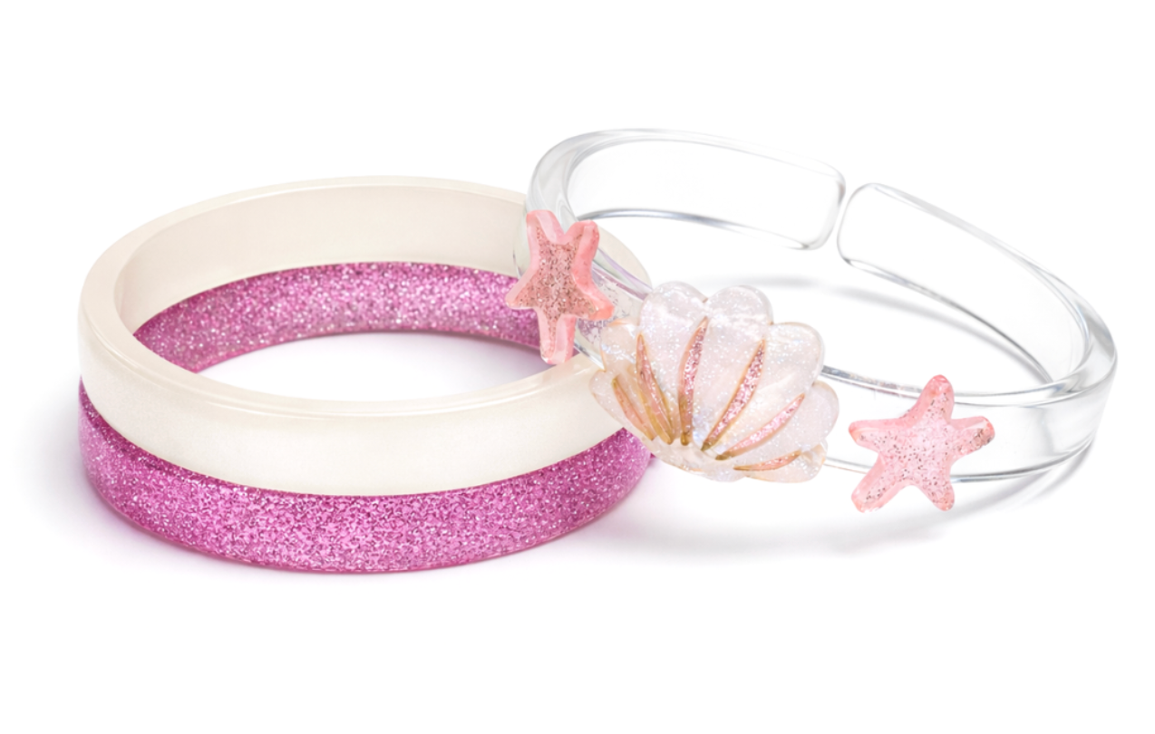 Lillies & Roses Seashell Pink Glitter Bangles features three handmade bracelets—one white, one pink glitter, and one clear adorned with pink stars and seashell decorations—that capture a playful beach spirit. Crafted in Brazil, this set is beautifully presented against a white background.