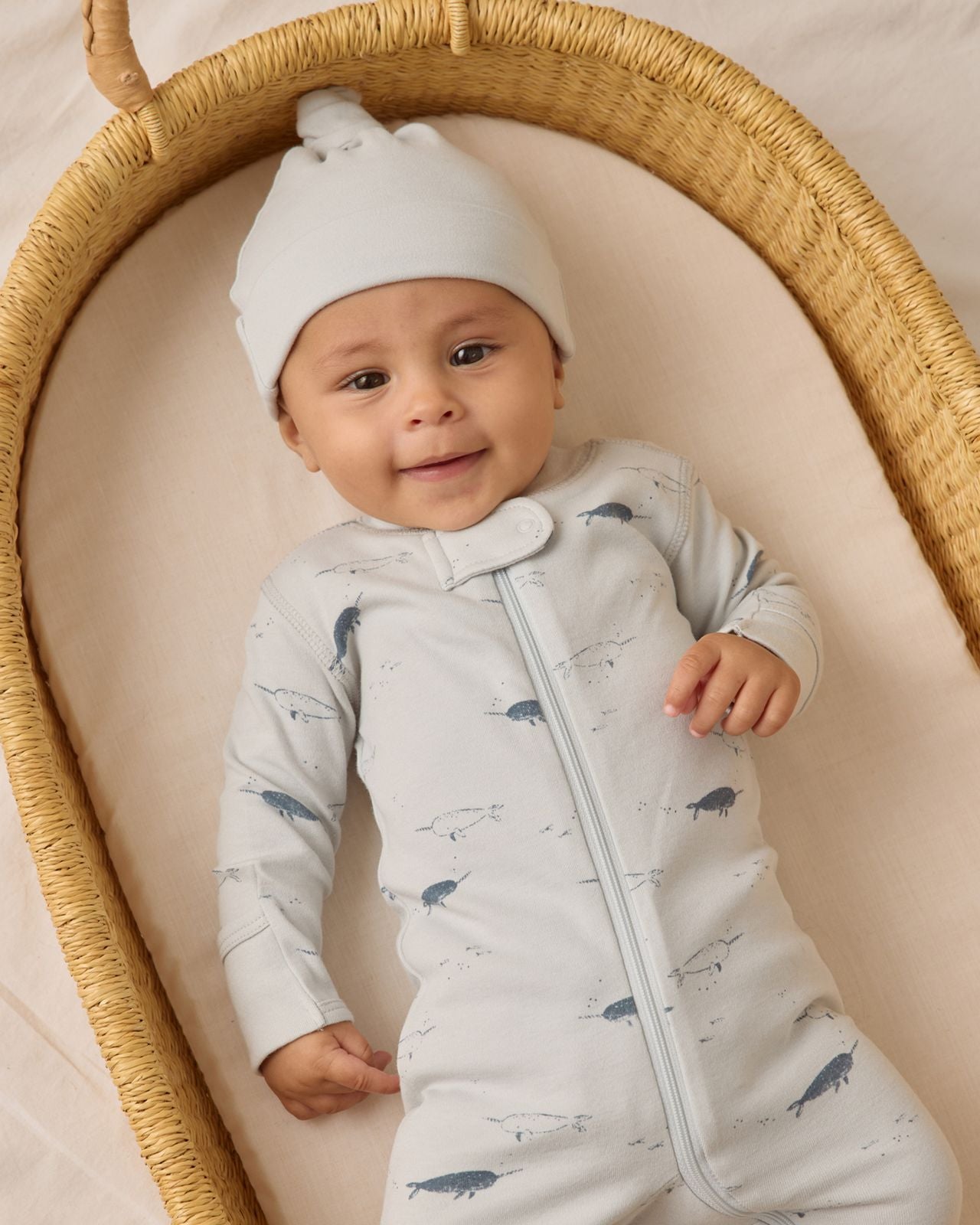 A baby in a Quincy Mae Baby Boys' Relaxed Fleece Jumpsuit lies in a woven bassinet, wearing a light blue hat and smiling gently.
