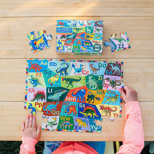 A child assembles the Eeboo Dinosaur Alphabet 20 Piece Puzzle on a wooden table, with the box and loose pieces nearby, making learning letters fun and interactive.