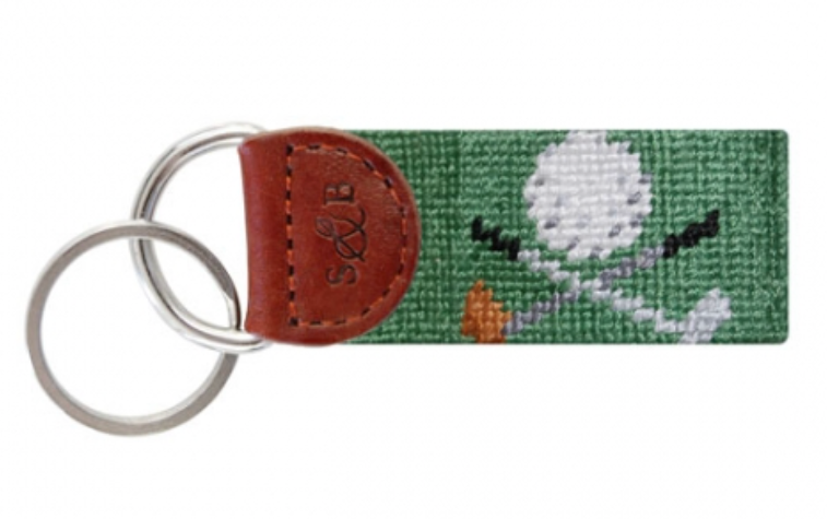 The Smathers & Branson Golf Clubs Sage Key Fob features a golf-themed design with crossed clubs and a ball on a green needlepoint loop. Its vibrant style is highlighted by a leather tab and two metal rings, making it an ideal accessory for any pocket or purse.