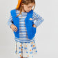 A young girl wears the Arsene & Les Pipelettes Boys' and Girls' Sailor Shirt, styled with a blue vest and printed skirt. She looks down with her hands in her vest pockets, her hair tied back with a bright orange scarf, standing against a plain light background.