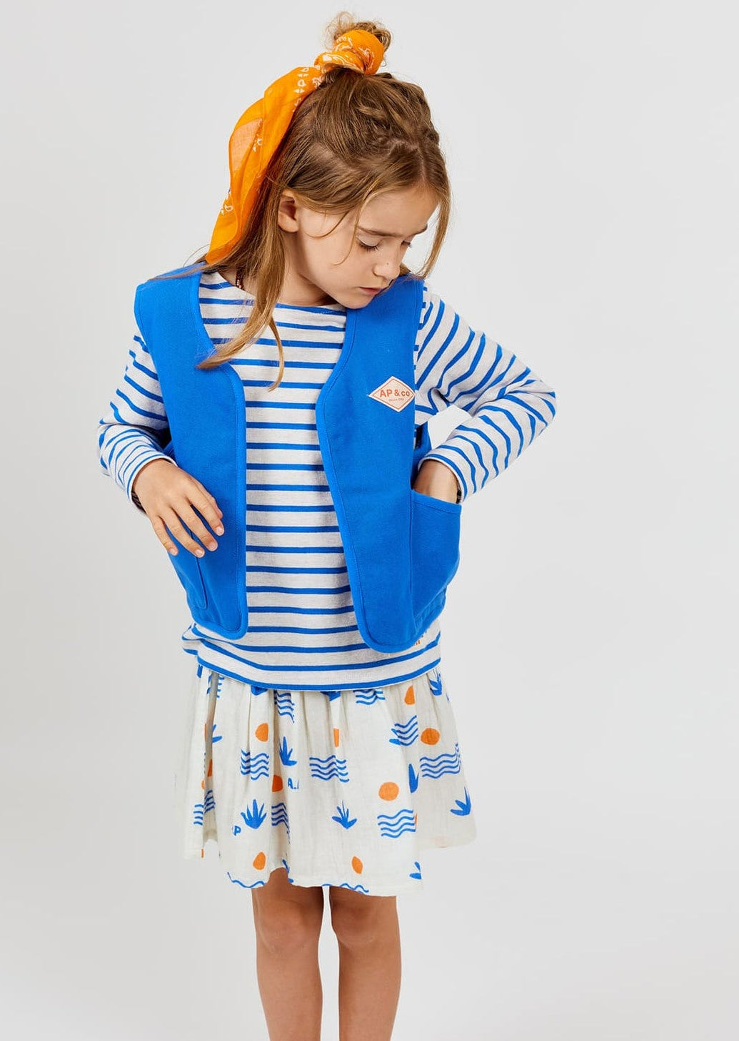A young girl wears the Arsene & Les Pipelettes Boys' and Girls' Sailor Shirt, styled with a blue vest and printed skirt. She looks down with her hands in her vest pockets, her hair tied back with a bright orange scarf, standing against a plain light background.