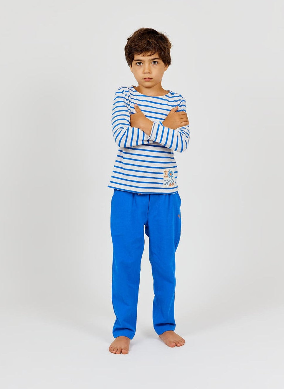 A young boy with short brown hair stands barefoot, arms crossed, wearing the Arsene & Les Pipelettes Boys' and Girls' Sailor Shirt—a nautical-inspired blue and white Breton striped top—paired with bright blue pants in soft organic cotton jersey against a plain light background.