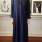 A person stands barefoot on a patterned rug wearing TWP's dark blue Jenny's Coated Linen Gown. In the background, two framed black and white photos adorn the wall.