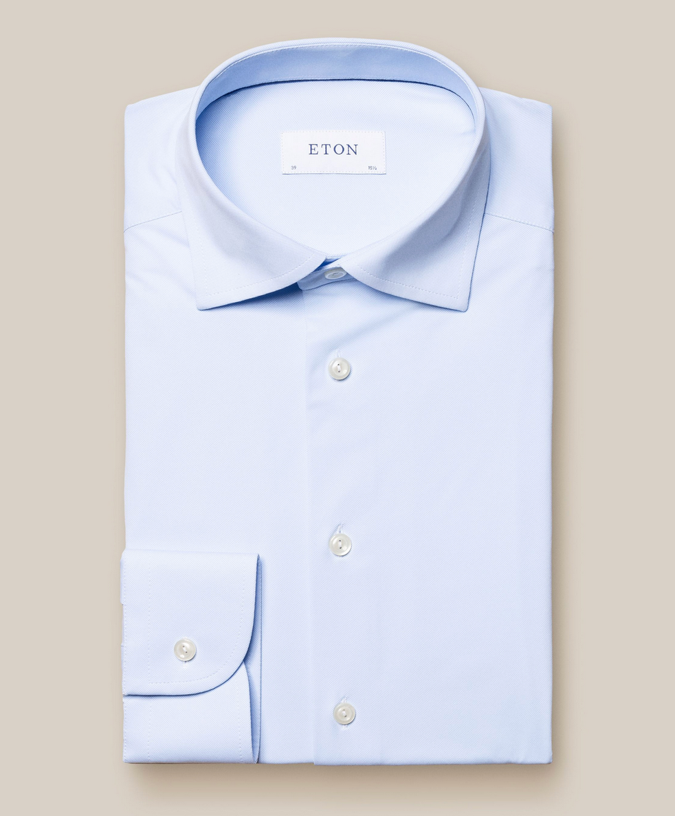 The Eton Four-Way Stretch Shirt, Slim Fit, in light blue features white buttons and a visible brand label, delivering tailored comfort on a neutral background.