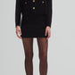 A woman wears the Blaze Milano Jealousy Shamo Bolero in black cotton velvet with gold-tone buttons, styled with a black mini skirt, sheer black tights, and high heels, posing against a plain background.