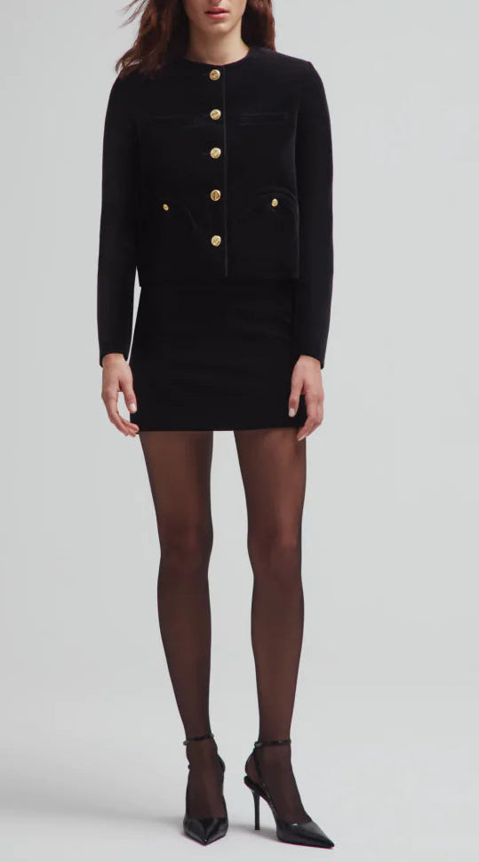 A woman wears the Blaze Milano Jealousy Shamo Bolero in black cotton velvet with gold-tone buttons, styled with a black mini skirt, sheer black tights, and high heels, posing against a plain background.