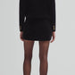 A woman faces away, wearing the Blaze Milano Jealousy Shamo Bolero in black cotton velvet with gold buttons, paired with a black mini skirt, sheer black tights, and black heeled sandals.