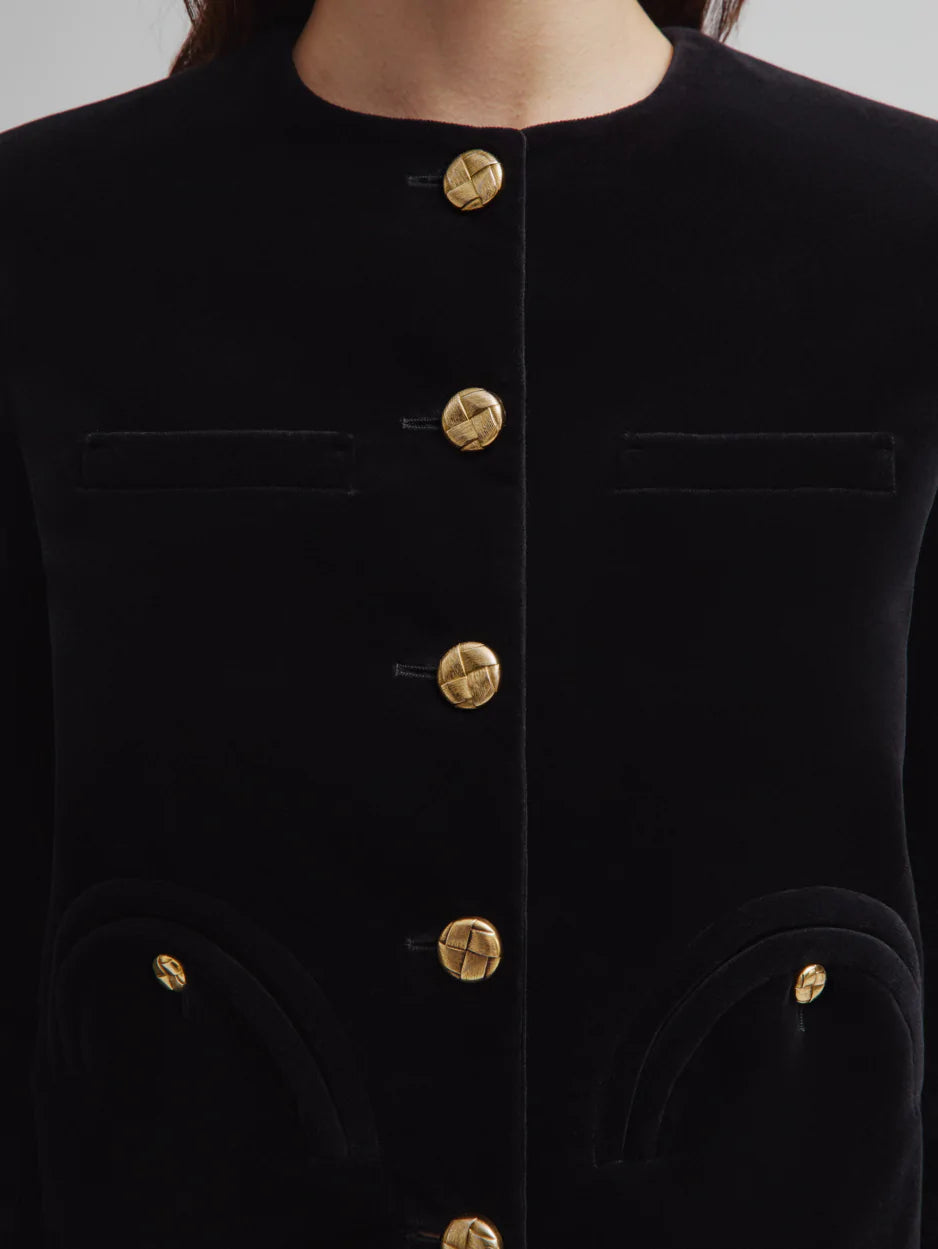 A close-up of the Jealousy Shamo Bolero by Blaze Milano, a black cotton velvet jacket with gold-toned buttons and four front pockets, worn by a person.