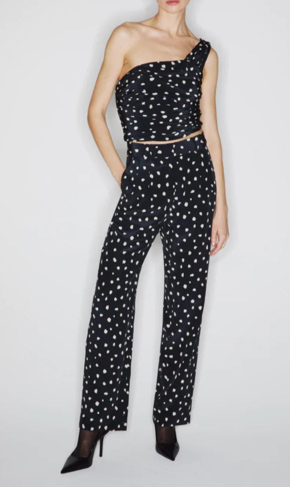 A woman models the Mariquita Nana Pants by Blaze Milano—high-waisted, straight-leg black pants with white polka dots—paired with a black one-shoulder sleeveless top and black high heels.