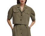 A person wearing the Blaze Milano Midday Sun Lola Jacket in olive green linen, featuring short sleeves, padded shoulders, two chest pockets, paired with matching high-waisted pants with pockets.