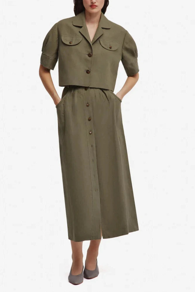 A woman wears the Blaze Milano Midday Sun Lola Jacket in olive green linen with padded shoulders and short sleeves, paired with a matching high-waisted A-line midi skirt; hands in pockets; gray shoes.