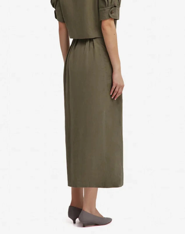A person stands facing away, wearing the Blaze Milano Midday Sun Nija Skirt in palm green and gray shoes, with arms relaxed by their sides against a plain white background.