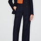 A person in Blaze Milano's First Class Fox Pants—dark navy, high-rise wool-mohair—paired with an orange blouse and light blue open-toe heels stands against a plain white background.