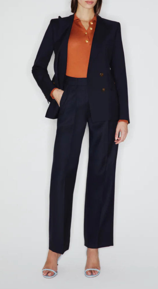 A person in Blaze Milano's First Class Fox Pants—dark navy, high-rise wool-mohair—paired with an orange blouse and light blue open-toe heels stands against a plain white background.