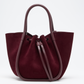 The Proenza Schouler Small Ruched Soft Suede Tote, in burgundy with leather handles and a drawstring closure, displays a sculptural silhouette against a plain white background.