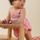 A baby wearing a Louise Misha Francesca Romper stands while holding onto a wooden chair.