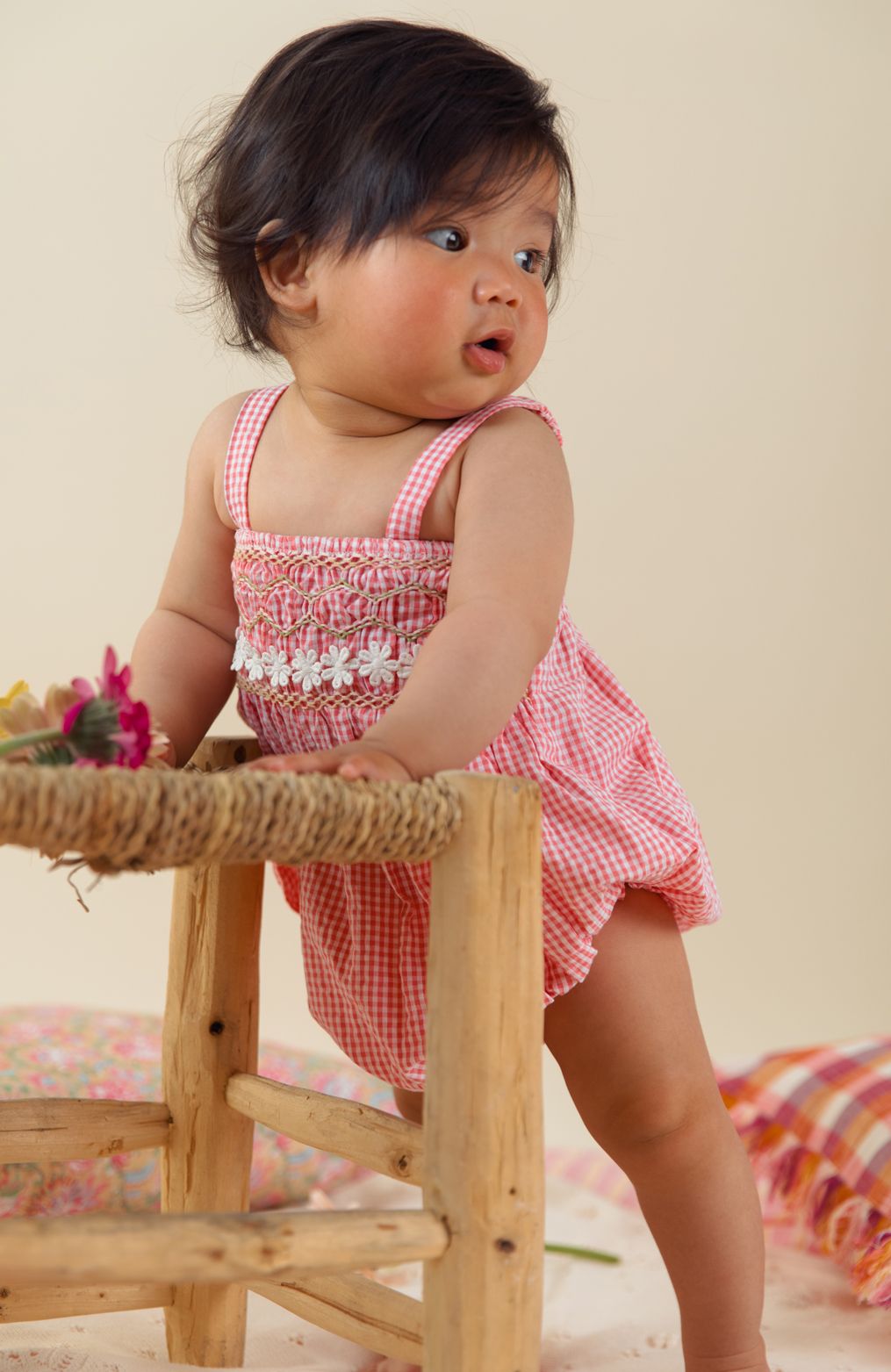 A baby wearing a Louise Misha Francesca Romper stands while holding onto a wooden chair.