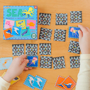 Two children play Eeboo’s Sea Little Square Memory Game by matching sea creature cards on a wooden table, with the colorful game box in view.
