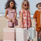 Three children standing on a wooden boardwalk by the beach with stylish Olli Ella See-Ya Suitcases, ready for vacation.