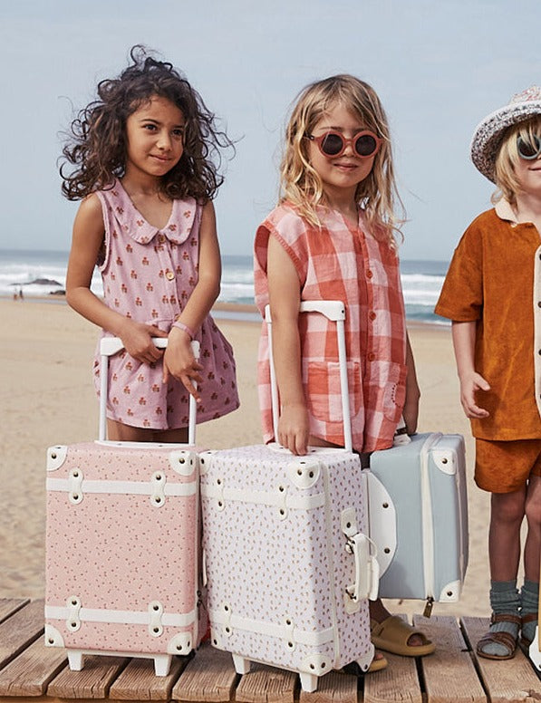 Three children standing on a wooden boardwalk by the beach with stylish Olli Ella See-Ya Suitcases, ready for vacation.