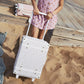 A child in a pink dress holding a white Olli Ella See-Ya Suitcase with a pattern, standing on wooden planks near a sandy beach.