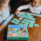 Two children sit at a wooden table playing the Eeboo Shiny Dinosaur Memory and Matching Game, with the game box and cards spread out before them, enjoying this educational kids’ game.