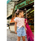 A young girl wearing a striped top and the Louis Louise Girls’ Andrea Denim Floral Short stands outside a store, holding a pink net bag and looking to the side.
